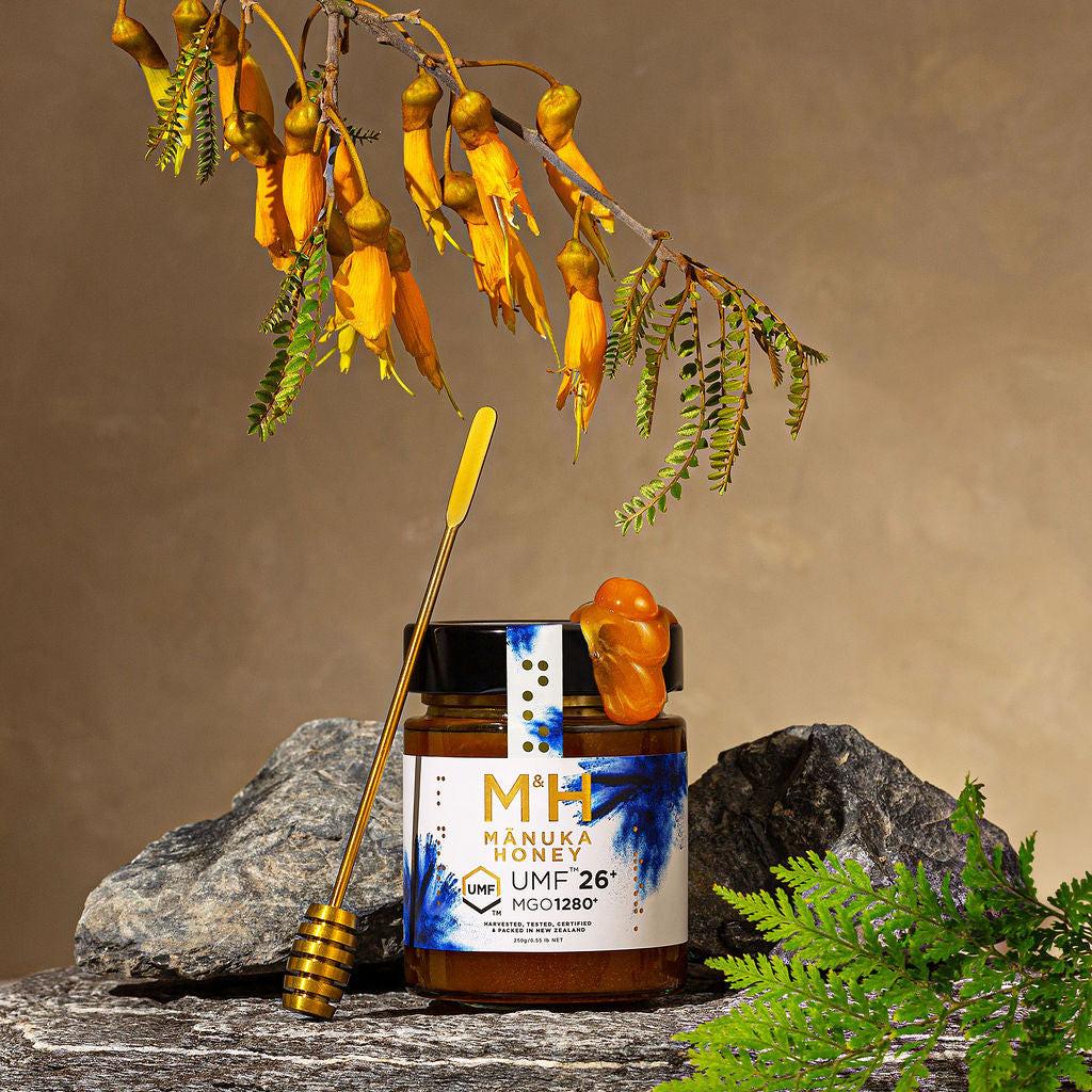 Manuka honey jar with a honey dipper on rocks, surrounded by flowers and plants.