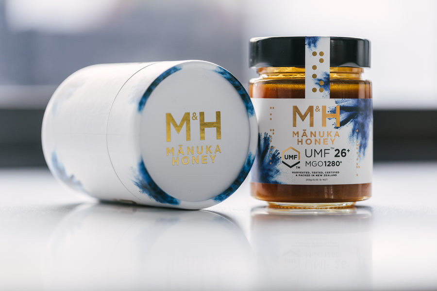 A jar of M&H Manuka Honey with a label indicating UMF 26+ and MGO 1280, suggesting a potent and high-quality honey product, next to gift packaging.