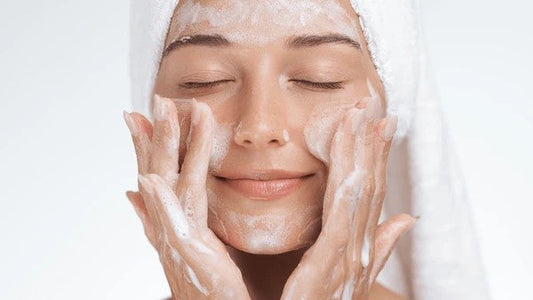 Serene looking young woman, massaging cream into her face, and with a towel wrapped around her head.