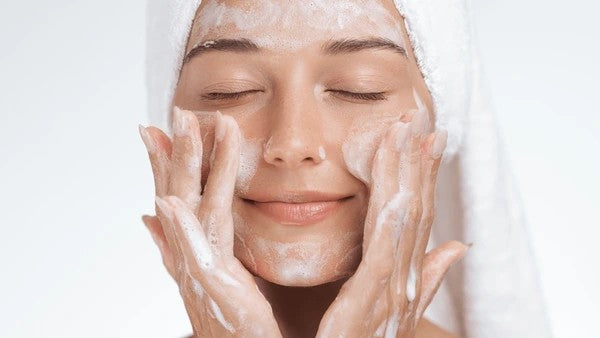 Serene looking young woman, massaging cream into her face, and with a towel wrapped around her head.