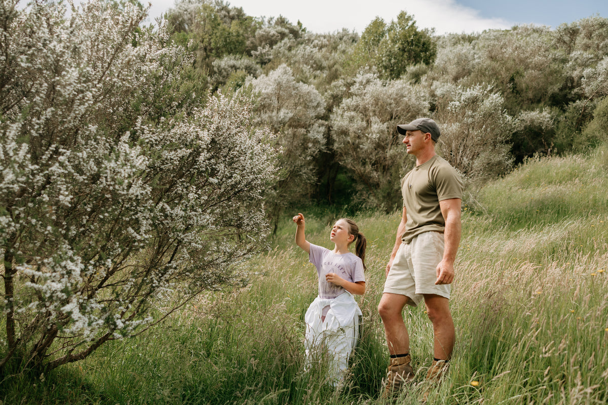 father and young daughter standing in a field with manuka honey trees blossoms and greenery.