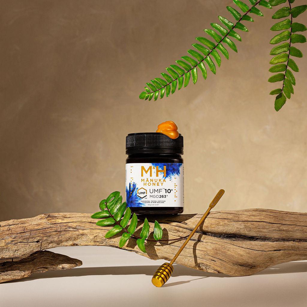 Jar of Manuka Honey with UMF 10+ label on a wooden surface with leaves and a honey dipper.